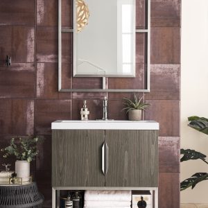 The modern bathroom showcases the Columbia 31.5 Bathroom Vanity in ash gray with brushed nickel, paired with a rectangular mirror and wood-paneled accent wall. Below the basin are open shelves holding rolled towels and a candle, complemented by plants, decor items, and a woven basket for a serene touch.