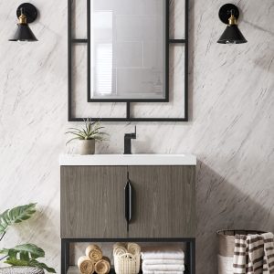 A modern bathroom showcases the Columbia 31.5 Bathroom Vanity in Ash Gray with Matte Black, paired with a black framed mirror and two black sconces. Rolled towels and a plant sit atop the vanity, while gray marble walls and herringbone floors complement a basket and stool near a leafy plant.
