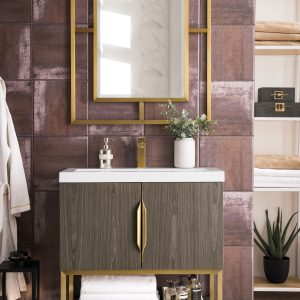 A modern bathroom boasts the Columbia 31.5 Bathroom Vanity in Ash Gray with Radiant Gold handles, complemented by a gold-framed rectangular mirror and shelves for towels and decor, all set against a textured brown wall and light wood flooring that elevate its elegance.