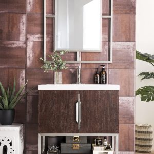 The Columbia 31.5 Coffee Oak Bathroom Vanity with a white countertop and brushed nickel accents is paired with a reflective mirror, small plants, and organized toiletries. A chevron pattern graces the floor, enhanced by a nearby white ceramic stool.