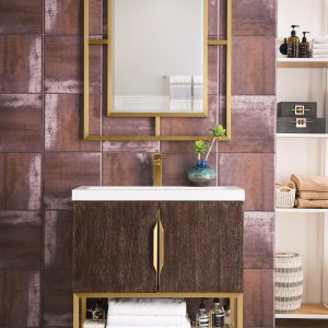 The Columbia 31.5 Bathroom Vanity in Coffee Oak with Radiant Gold features a gold-framed rectangular mirror, textured brown tiled wall backdrop, and a brown oak cabinet with gold accents. Shelves hold white towels and toiletries, while a small green plant adorns the countertop.