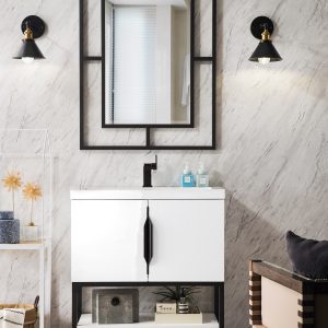 A modern bathroom showcases the Columbia 31.5 Bathroom Vanity in glossy white and matte black, paired with a marble wall and black-framed mirror flanked by two black sconces with gold accents. A light wooden herringbone floor and decorative shelves provide warmth on the left.