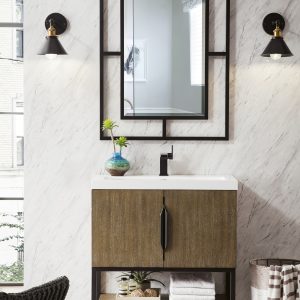 A modern bathroom features the Columbia 31.5 Bathroom Vanity in Latte Oak with a wood-textured cabinet, undermount sink, and matte black faucet. A rectangular metal-framed mirror hangs above, flanked by black and gold sconces, with marble-patterned walls and a small potted plant enhancing the decor.