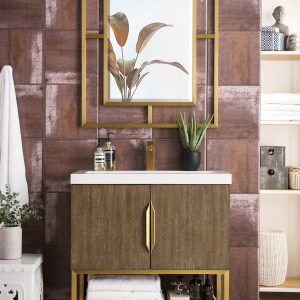 An elegant bathroom features the Columbia 31.5 Bathroom Vanity in Latte Oak and Radiant Gold. A leafy plant and soap dispenser adorn the counter, while a large gold-framed decorative mirror hangs above. Towels and decor items are neatly arranged on adjacent shelves.