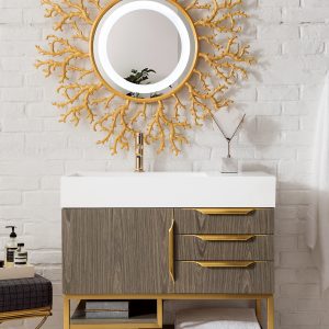 A stylish bathroom showcases the Columbia 36 Single Bathroom Vanity in Ash Gray with Radiant Gold accents. An open shelf holds folded towels, and a round mirror with a gold coral-like frame hangs on the white brick wall above the sink, enhanced by a chair and soap bottle for added charm.