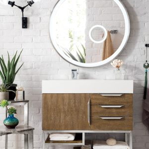 A modern bathroom showcases a Columbia 36 Single Bathroom Vanity in Latte Oak with Brushed Nickel hardware. The vanity features drawers and open shelves for towels and decor. White brick walls, a small table with plants, an illuminated round mirror, and wall-mounted light complete the contemporary design.