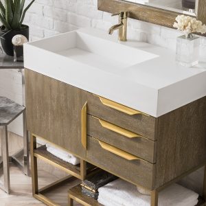 The Columbia 36 Single Bathroom Vanity in Latte Oak features a white countertop, gold faucet, and wooden cabinet with radiant gold handles. Towels neatly rest on an open lower shelf while a potted plant and decorative item adorn the space beside a large mirror above the sink.