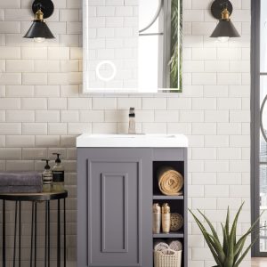 A modern bathroom features the Alicante 24 Bathroom Vanity in Grey Smoke with a white sink, open shelving, and a square mirror above. Two black sconces flank the mirror, while a potted plant sits next to a small black side table for towels and toiletries.