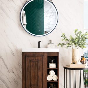 The Alicante 24 Bathroom Vanity in Mid Century Acacia and Matte Black is complemented by a round mirror above its wooden cabinet, which features shelves for towels and toiletries. Nearby is a small table with plants and bottles, all set against a brightly lit marble wall and light wood floor.