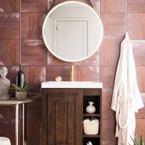 A modern bathroom flaunts the Alicante 24 Bathroom Vanity in Mid Century Acacia with Radiant Gold, crowned by a round mirror. Decorative items on shelves, a white robe on the textured brown wall, a small side table, and a plant enhance the light herringbone-patterned floors elegance.