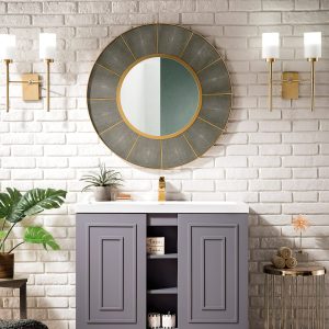 The elegant bathroom design highlights the Alicante 39.5 Bathroom Vanity in Grey Smoke with Radiant Gold accents, complemented by a round gold-framed mirror and modern wall sconces. A white brick wall and herringbone-patterned floor are adorned with a potted plant and decorative items.