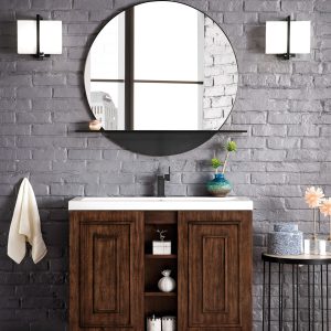 A modern bathroom features the Alicante 39.5 Bathroom Vanity in Mid Century Acacia and Matte Black, with a round mirror and two wall sconces above. It has a white countertop, open shelves with decor, and is paired with a black metal side table with flowers, enhancing the brick walls and wooden floor.