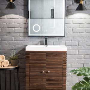 A modern bathroom features a Linden 24 inch Bathroom Vanity in Mid Century Walnut with a white sink against a gray brick wall. Above, a backlit mirror is flanked by two black wall-mounted lights. A small table nearby holds rolled towels and a green plant.