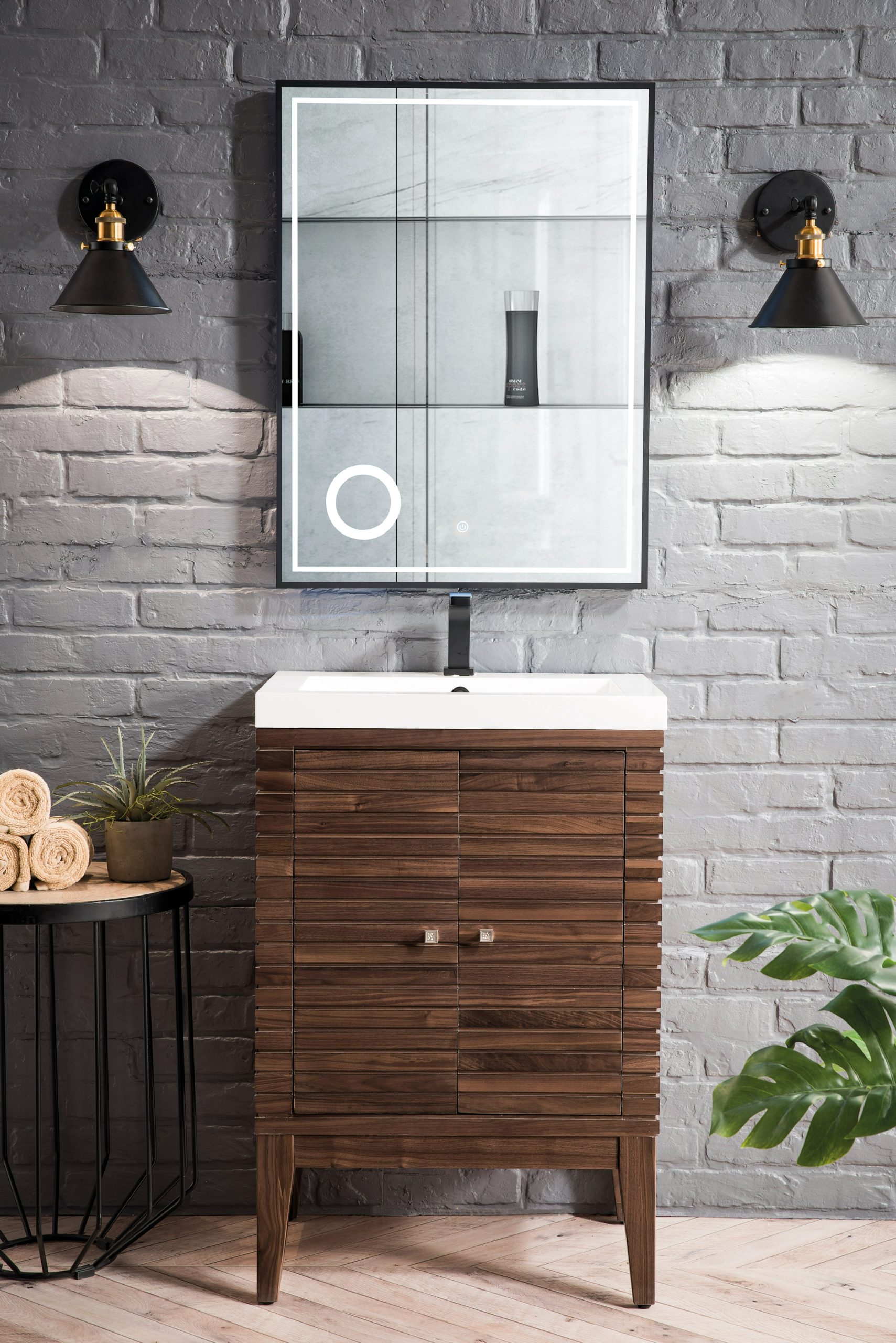 A modern bathroom features a Linden 24 inch Bathroom Vanity in Mid Century Walnut with a white sink against a gray brick wall. Above, a backlit mirror is flanked by two black wall-mounted lights. A small table nearby holds rolled towels and a green plant.