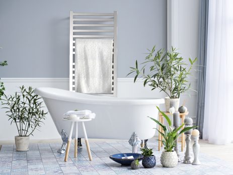 A modern bathroom features a white freestanding tub, green plants, and a rug. Towels rest on a small stool, while a towel hangs on the Quadro Q2142 16 Bar Hardwired Towel Warmer in Polished against a light gray wall bathed in natural light.
