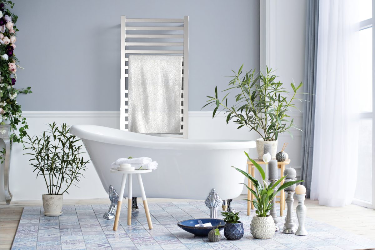 A modern bathroom features a white freestanding tub, green plants, and a rug. Towels rest on a small stool, while a towel hangs on the Quadro Q2142 16 Bar Hardwired Towel Warmer in Polished against a light gray wall bathed in natural light.