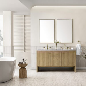 A modern bathroom showcases a Hudson 60 inch Double Vanity Cabinet in Light Natural Oak with gold fixtures and dual sinks. Above it hangs rectangular mirrors. Nearby, a white bathtub and wooden side table with essentials enhance the light-colored tiles, framed by a large window with shutters.