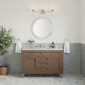 A modern bathroom highlights the Chicago 48-inch Single Bathroom Vanity Cabinet with a marble top and round mirror. Three lights with clear glass bulbs stylishly illuminate above it, while light gray subway tiles cover the lower wall. A tall plant enhances the space near the vanity.