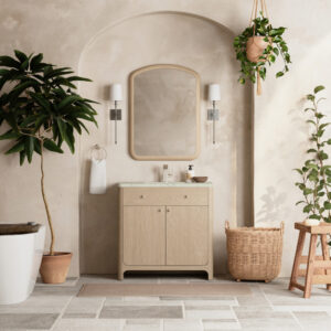 A minimalist bathroom showcases the Gracyn 36 inch Bathroom Vanity with a round-edged mirror and two wall-mounted lights. Potted plants, a wicker basket, and a wooden stool add natural touches, while large, irregular stone tiles cover the floor.