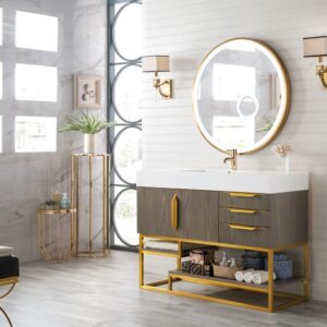A modern bathroom showcases the Columbia 48 Inch Single Vanity Cabinet with gold accents, paired with a round gold-framed mirror, tidy towels, plants, and decor against light walls and expansive geometric windows.