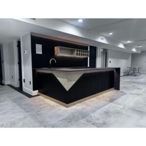 Modern basement bar with a geometric black and light wood design, under-cabinet lighting, open shelving, a gold faucet, and a tiled floor. A ping pong table is visible in the background.