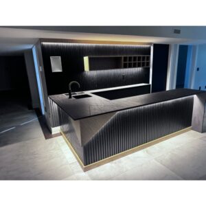 Modern kitchen with a black minimalist island, integrated sink, and under-cabinet lighting. Vertical wood paneling and gold accent base highlight the sleek, contemporary design. Light gray floor tiles complete the space.